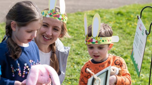 Two young children with woman, wearing bunny ears at an Easter activity point with grass in background